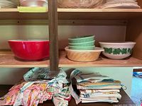Vintage Pyrex mixing bowls displayed on a shelf, including red, beige swirl, green, and white bowls with ivy pattern alongside folded fabric items below shelf.