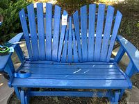 Front view of blue Adirondack style double glider chair showing slatted seat and backrest, armrests and gliding base on stone paver patio with landscaping.