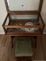 Photo showing wooden display case with glass top open, small green upholstered stool in front, items inside display case including glass rods, candies, glass dish, and doily visible.