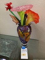 Front view of the purple vase with multiple colorful glass flowers arranged inside including red, pink, and orange flowers with green stems and a brown leaf.
