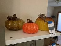 Three hand-blown glass pumpkins displayed on a white shelf in natural indoor lighting, showing their colors and stems clearly.