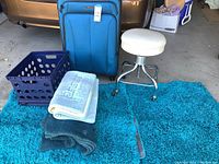 Photo showing the full lot of blue suitcase, teal area rug, two folded blankets, mid-century white rolling stool, and purple plastic bin