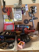 Workbench with assorted tools and materials, pegboard with hanging hand tools and accessories, Porter-Cable cordless drill with charger, smoke alarm package, multi-surface sandpaper, various hardware containers and baskets on surface.