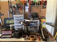 Wide view of pegboard with screwdrivers, pliers, wrenches, and shelves with storage drawers and assorted tools on table including power drill, charger, and hand sander.