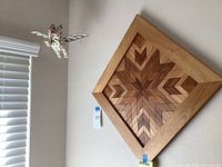 Photo shows square wooden wall art with geometric inlay pattern hanging on wall with flying cat sculpture suspended nearby.
