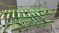 Full view of the green wrought iron patio lounger showing slatted seat, decorative circular and scroll metalwork on armrests and backrest, and absence of wheels.