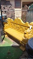 Front side of ornate yellow painted cement bench with decorative patterns and animal head armrests next to chain link fence.