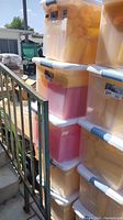 Five clear plastic tubs with white lids stacked outdoors. Containers are filled with numerous yellow and red condiment squeeze bottles.