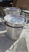 Photo showing perforated metal pot with handles and clear lid wrapped in plastic on top, with fryer baskets inside and beside it