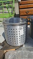 Large aluminum fryer pot with perforations on sides, no lid, metal handle on rim, requires cleaning.