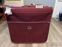 Front view of burgundy garment carrier luggage showing large zippered front pocket and leather handle with tags