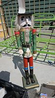 Front view of tall wooden nutcracker statue with green jacket, red pants, black boots, black hat, and white beard. The figure stands on a base and is shown outdoors on a green bench.