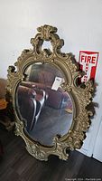 Full view of a large vintage ornate oval mirror leaning against a wall, showing the overall size and look including part of the surrounding area.