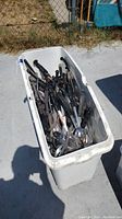 Top down view of white plastic bin filled with various metal slotted spoons and ladles with black handles.