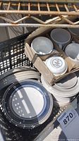 Photo showing stacked off-white and blue striped stoneware plates with rustic finish, along with cardboard boxed white china cups and bowls below a metal rack.