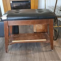 Front view of wooden piano bench with black leather cushioned top showing scratches and scuffs on wood and missing button on leather.