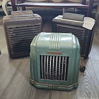 Three vintage small metal heaters, including one green Thermador and two dark metal heaters with horizontal slats, visible heavy rust and wear.
