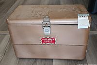 Front view of vintage Little Brown Chest metal ice cooler showing red and white logo, latch, and brown worn finish.
