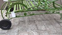 Side view of vintage green-painted wrought iron lounger showing decorative scrollwork, slatted seating area, and black rubber wheel attached.