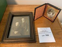 Framed black and white photo of an older woman and man beside an antique daguerreotype in its open case on a wooden table.