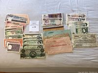 Top-down photo showing a spread of various paper currency notes from Commonwealth countries arranged in rows and stacks. Visible notes include Canadian, Indian, Bahamian, Australian, and English bills along with promissory notes in pastel colors.