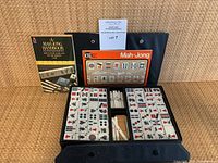 Black folding case open showing organized mahjong tiles, scoring sticks, and two books propped behind
