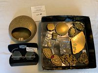 Overview photo of lot showing all brass items arranged in box next to embossed brass compact or ashtray dish and eyeglasses in case