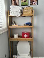 Bamboo shelving unit standing over a toilet in a bathroom, holding towels, a basket, cosmetic items, a first aid box, and toilet paper.