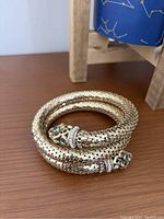 Photo showing coiled gold-tone snake bracelet with emerald green eyes and rhinestone accents, placed on wooden surface.