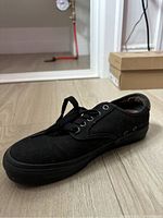 Single black Vans skate shoe on wood floor with shoe boxes in background.