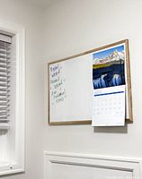 Photo of large whiteboard mounted on wall with natural wood frame. Writing in different colors and a calendar hanging on it.