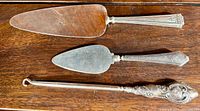 Two antique cake servers laid side by side showing wide and narrow blades with silver handles.