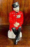 Front view showing Chelsea Pensioner sitting with hands clasped, wearing red coat and black hat with 'R H' lettering.