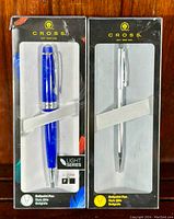 Two new CROSS pens in original black packaging, one blue from the Light Series with 0.8mm tip and one silver plain pen, displayed side-by-side showing full front views of boxes with branding.