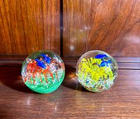 Two hand blown glass paperweights featuring jellyfish shapes in red and yellow with blue highlights, set on a wooden surface.