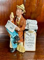 Front view of the Royal Doulton "Stop Press" figurine showing the seated man holding a newspaper and sandwich, sitting on a crate with printed text.