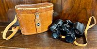 Vintage Kurt Muller binoculars displayed next to original tan leather carrying case with shoulder strap on wooden surface.