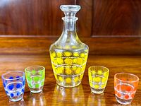 Front view of glass decanter and four matching colorful shot glasses arranged in a row.