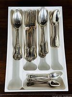 Assortment of silverplated spoons and forks arranged in a white compartment tray showing two distinct but similar ornate handle patterns.