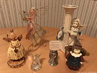 Seven angel-themed figurines and one candle holder grouped on a table with wallpaper background