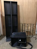 Black tall CD tower, metal stand, and fabric carrying case arranged on carpet in front of wooden door.