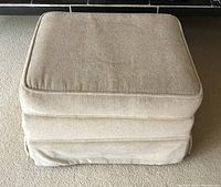 Front facing image of a beige chenille square ottoman on rollers, showing the full item with a cushioned top and base, placed on carpet.