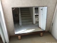 Rectangular dresser mirror with clear reflective surface and wooden legs, placed on floor against wall near a doorway and staircase.