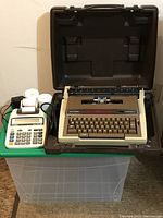 Photo showing both the Smith-Corona electric typewriter inside its carrying case and the Canon adding machine positioned on green table surface, highlighting the full lot contents including accessories such as power cables and paper rolls.