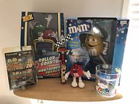 Photo showing all lot items together on a shelf: M&M's roller coaster dispenser box, Tony Stewart Matchbox car, shower radio, hand fan, key chain, glass cup, and a small wooden totem figurine.