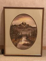 Framed oval landscape painting of mountain and river with trees under moody sky.