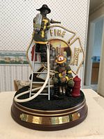 Front and side view of firefighter figurine titled 'Out of Harm's Way' showing two firefighters in detailed uniform, one on ladder with hose, one seated with helmet next to fire hydrant, mounted on wooden base.