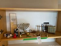 Wooden cabinet shelf showing lot items including brass Menorah, apple paperweight, brass picture stand, candle snuffers, candle shaver, packaged candles, tin cups and small brass items
