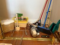 Overview of lot showing brooms, cleaning product caddy, handheld vacuum, dish rack, toilet brush, compost bin and box of bags.
