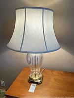 Front view of the lit lamp showing clear glass body, brass base, and white fabric shade.